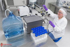 Scientist in lab coat, gloves and hair net operates laboratory equipment, beakers in foreground