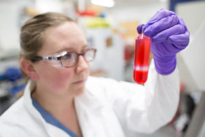 Female researcher in gloves, lab coat and safety glasses, holds up a red-filled vial for inspection