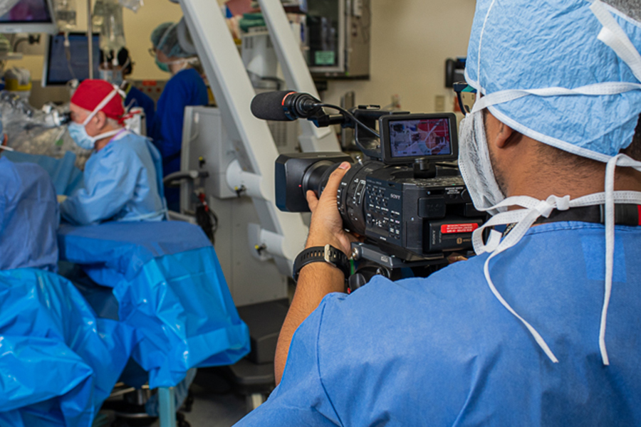 Videographer Pathum at work in scrubs. Camera focused on surgeons in background