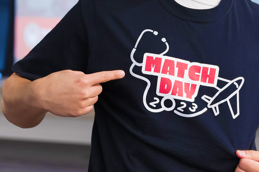 Photo of hand pointing to logo on black t-shirt. Logo is red and white. Text reads Match Day 2023 with stethoscope and airplane graphics